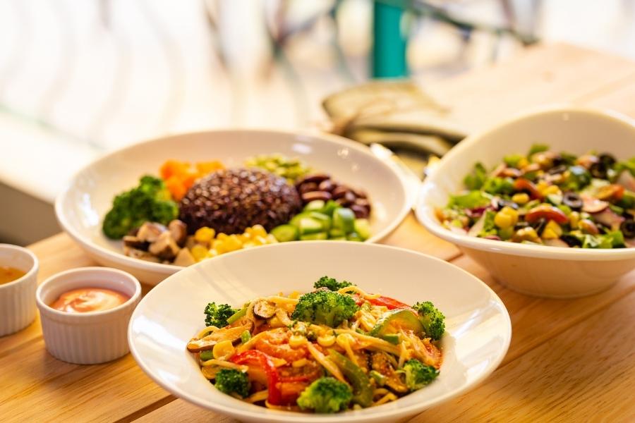 Three white bowls filled with colorful healthy meals including vegetables, grains, and legumes on wooden table