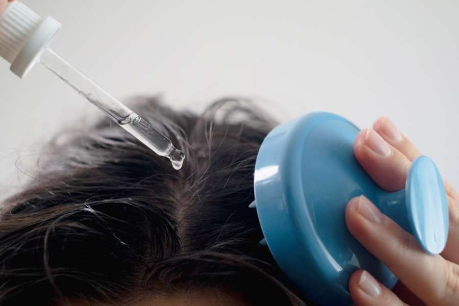 Hand applying scalp oil with a dropper while using a blue scalp massage tool.