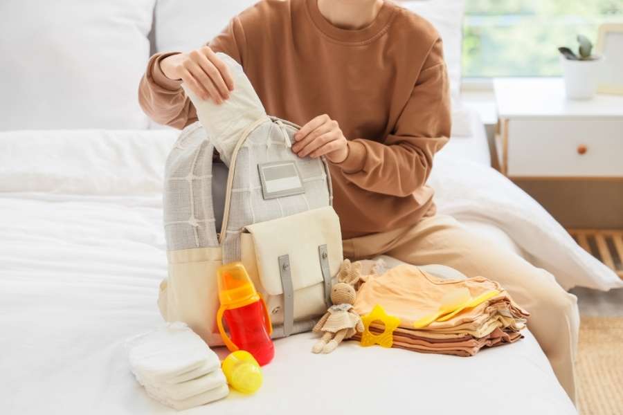 A person sitting on a bed while organizing items into an emergency diaper bag surrounded by baby clothes and bottles.